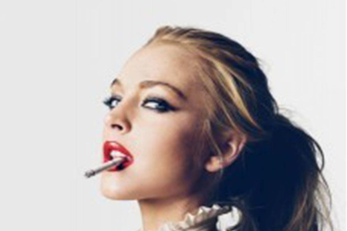 A large photo of a young blonde person with smoky eyes who holds a burning cigarette between their red lips