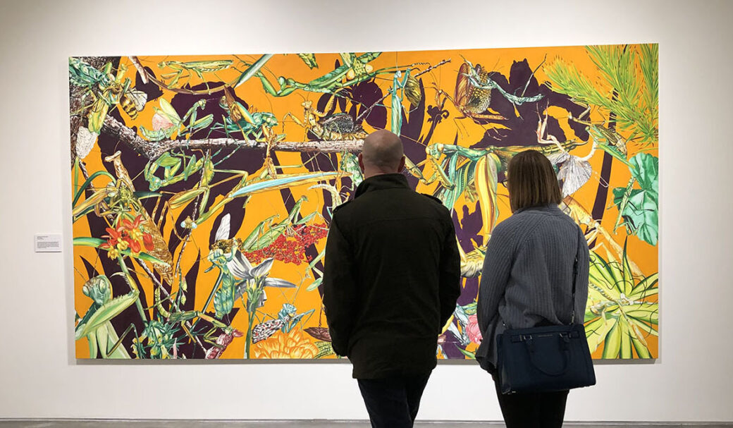 Two people view a brightly colored painting featuring mantises and flowers