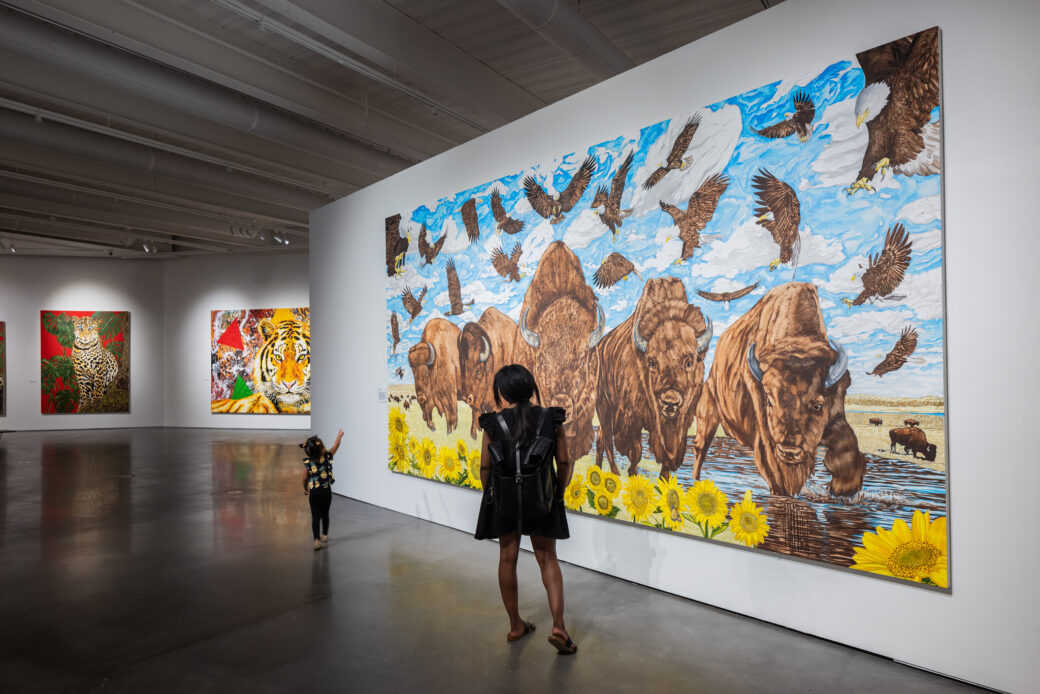 An adult and child stand in front of a large painting featuring bison and eagles. The child points at the painting. Two additional paintings featuring a leopard and a tiger are visible in the background.