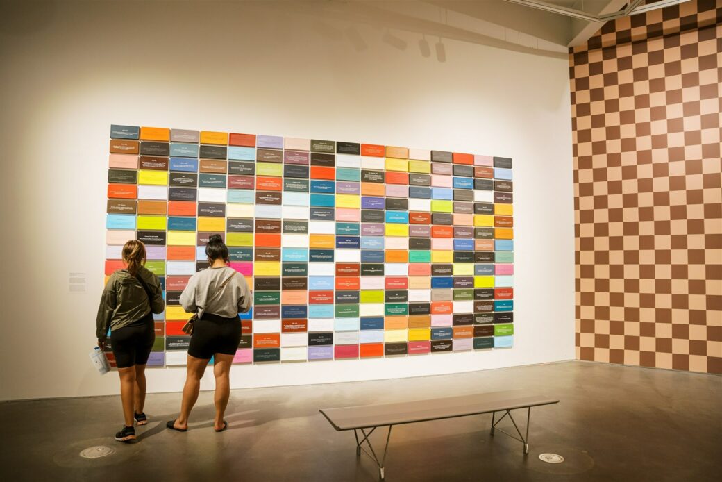 Two viewers stand in front of a wall of small, colorful canvases. A checkerboard wall is visible to the right.
