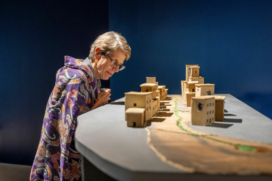 A person with short hair and glasses looks closely at a row of miniature beige buildings