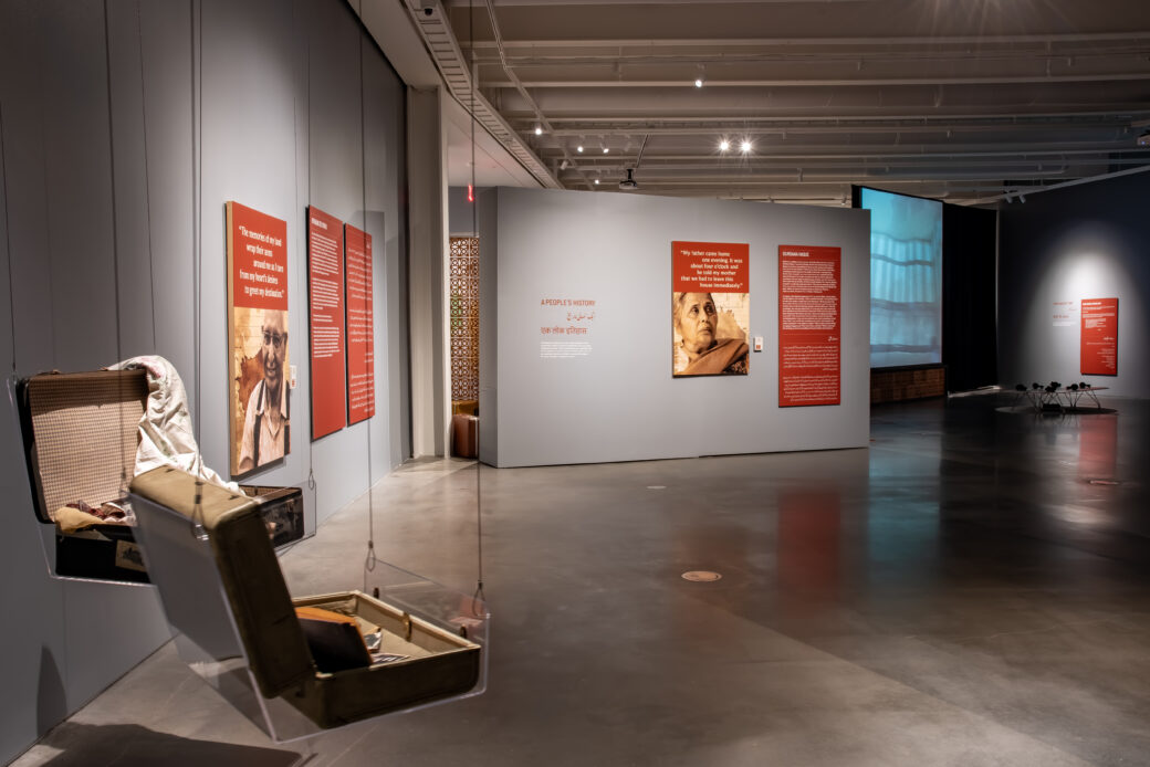 A gallery with suitcases suspended from the ceiling and red panels with text and pictures hanging on the walls