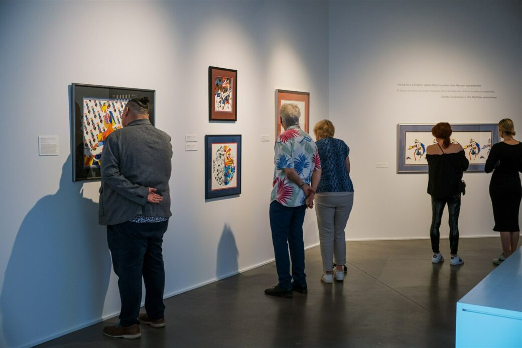 Five people stand in an art gallery with white walls and a cement floor. On the walls, we can see five prints framed and hung up. The prints are lively with vibrant colors and patterns.