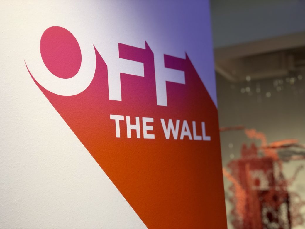 Close-up Off the Wall logo with Sarah Ahmad's work in the background