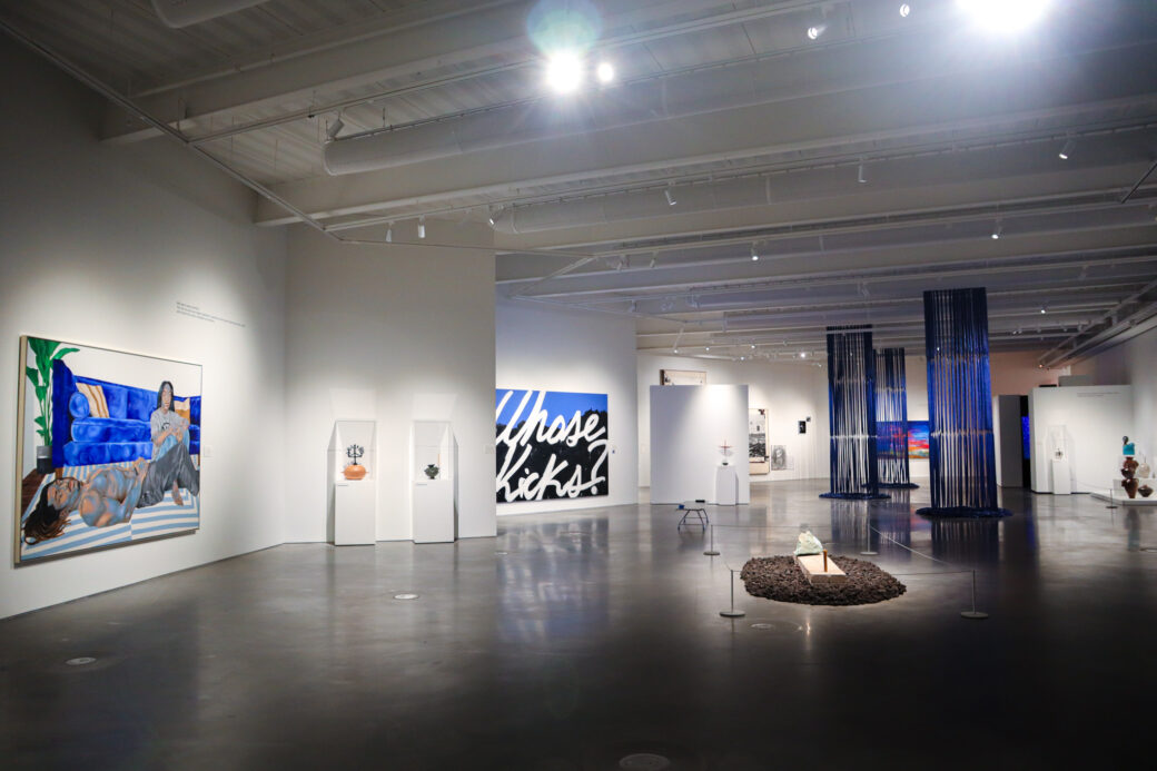 A large painting of a couple hangs on the left side of an art gallery. Three large columns of blue tarp tendrils hang in the center. Ceramic vessels line each side. To the back left we can see a large painting in white font that reads WHOSE KICKS?