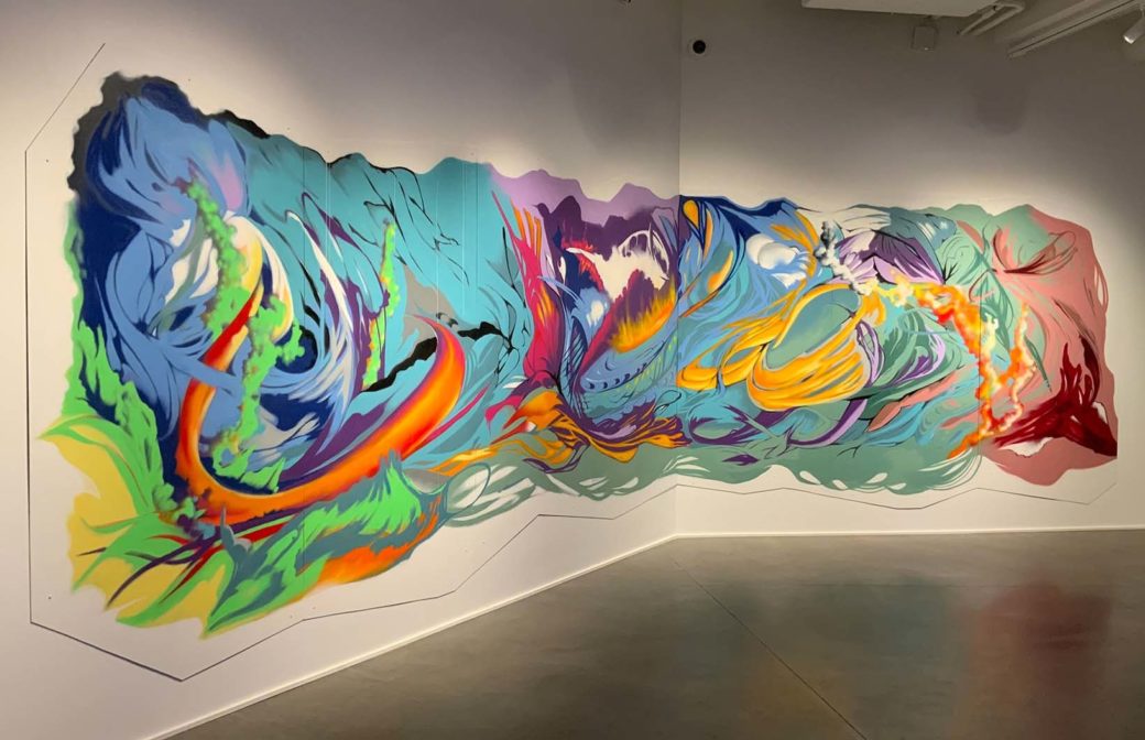 An abstract spray paint mural with intermingling warm and cool neon tones in broad strokes across multiple walls in a white-walled gallery with concrete floors