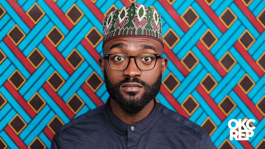 A person with a black beard and surprised expression wears black-framed glasses and a green and red patterned flat-top hat in front of a backdrop painted with a woven blue and red diamond pattern