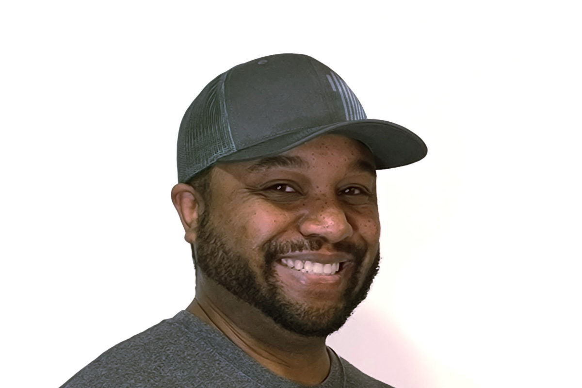 Man with facial hair and a grey baseball cap smiles at the camera against a white background.