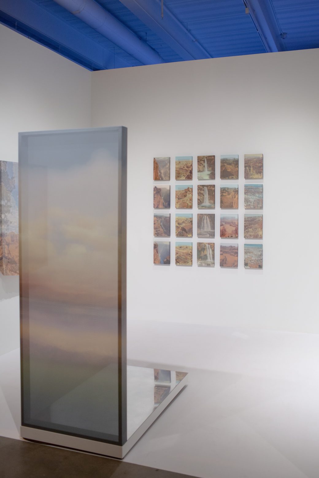 A vertical silk canvas stands in front of a wall with 20 small landscapes hung on it in a grid