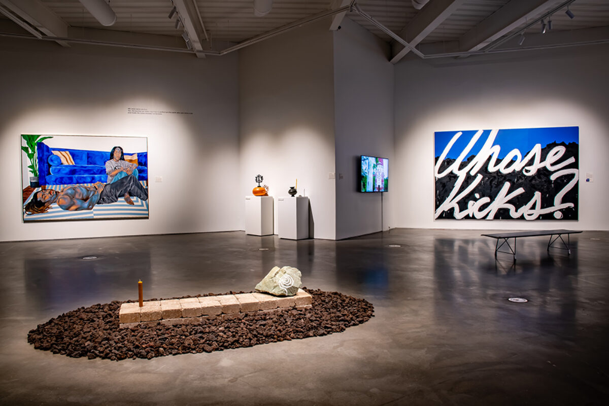 A stone rectangle holding a painted rock and a beeswax candle sits in the foreground. In the background, two large paintings, two vases on plinths and a video screen are visible.
