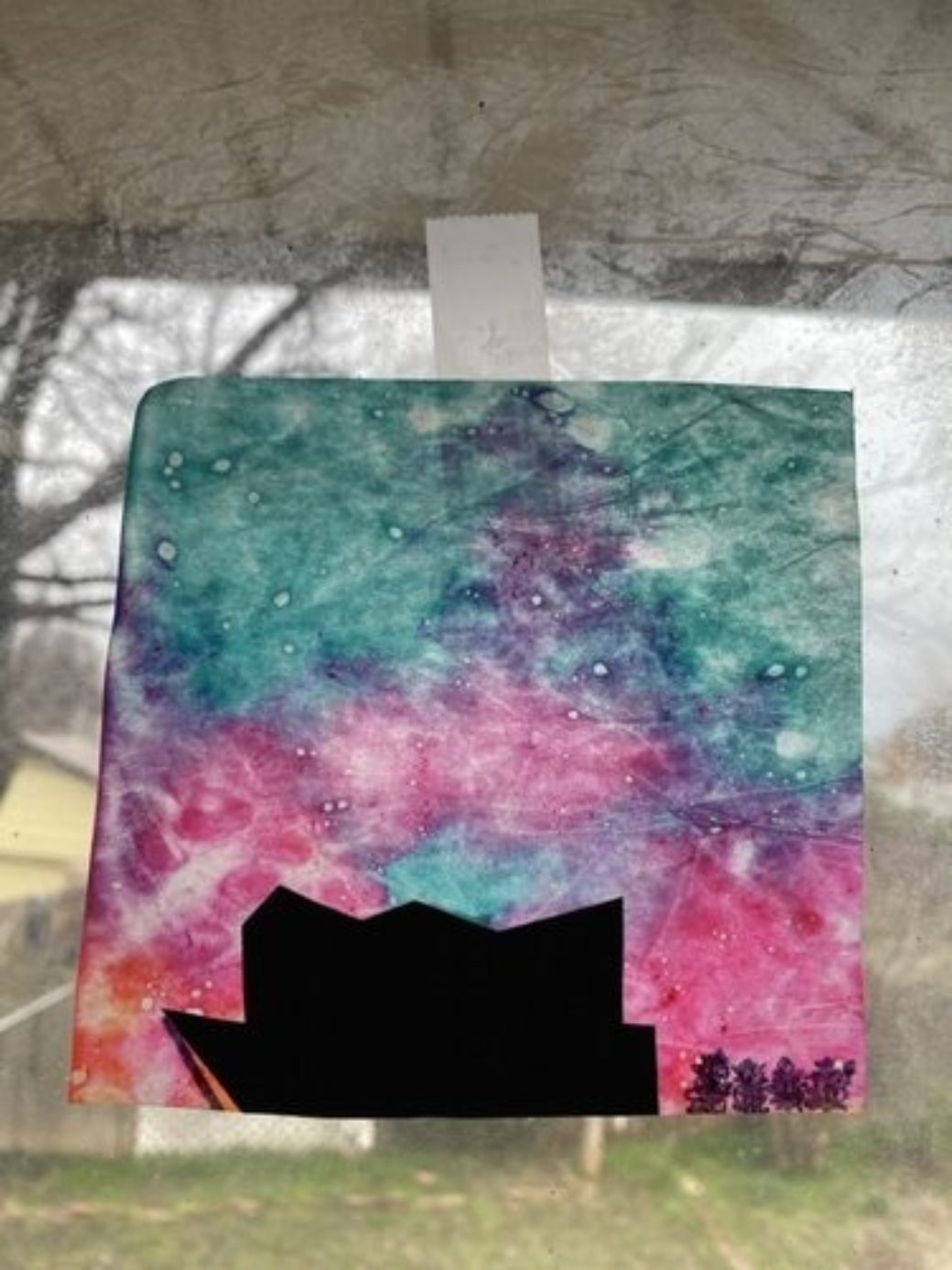 A multicolored, tie-dyed wax paper featuring the silhouette of a building drawn on it is taped in a window