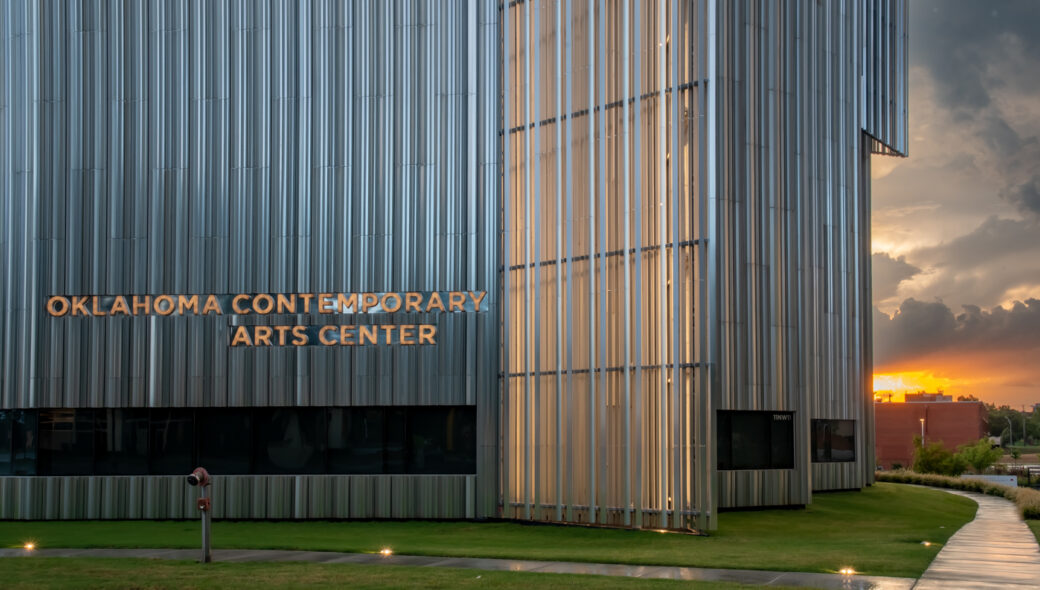 A silver building with words on the side reading "Oklahoma Contemporary Arts Center" and a sunrise in the background