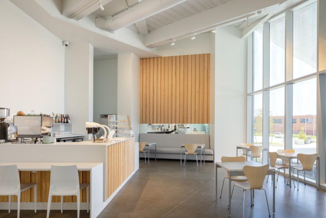 A cafe with white walls, wooden panels and large windows stands empty
