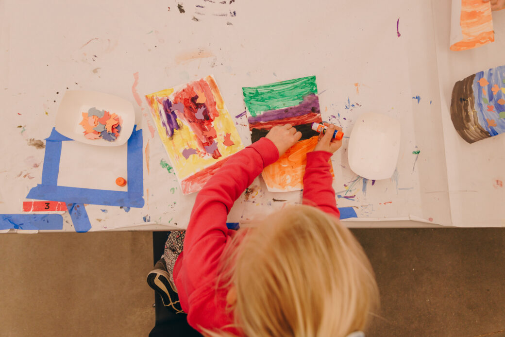 A child with blonde air and a pink, long sleeved shirt is working on an art project, using a glue stick on a colorfully drawn paper.