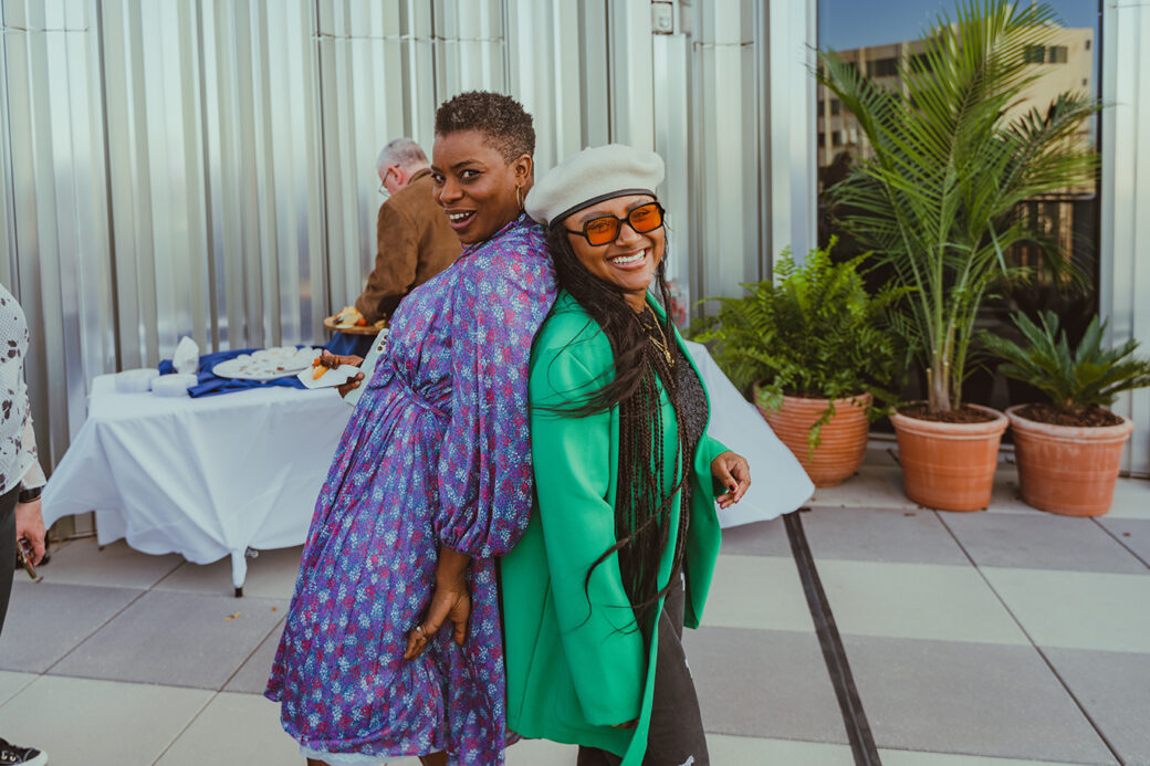 A short-haired Black woman in a blue and purple dress stand back-to-back with a Black woman wearing braids, a beret, sunglasses and a green jacket