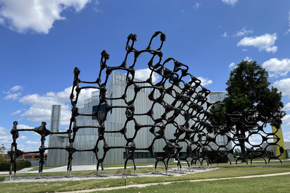 A giant sculpture of used tires twisted onto metal frames sits in a green park in front of a silver building