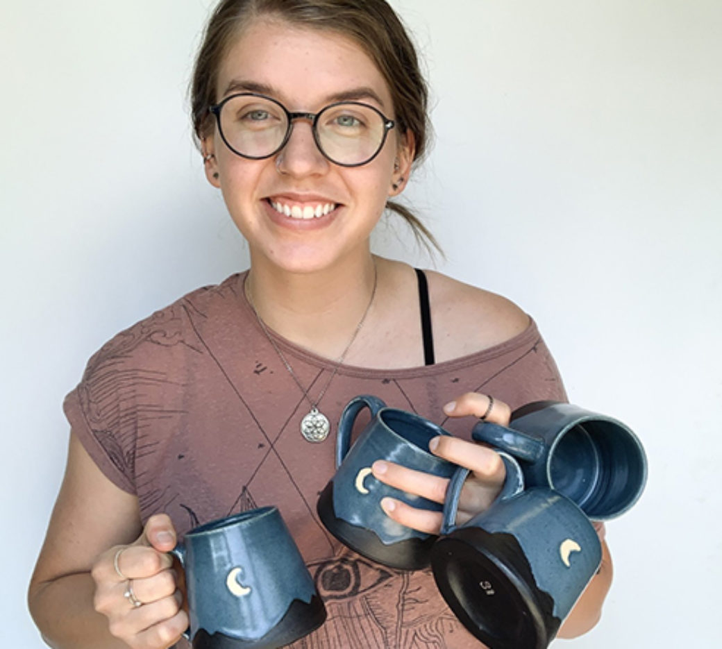 Color photo of a woman with glasses wearing an off-the-shoulder t-shirt holds four ceramic mugs smiles at the camera.