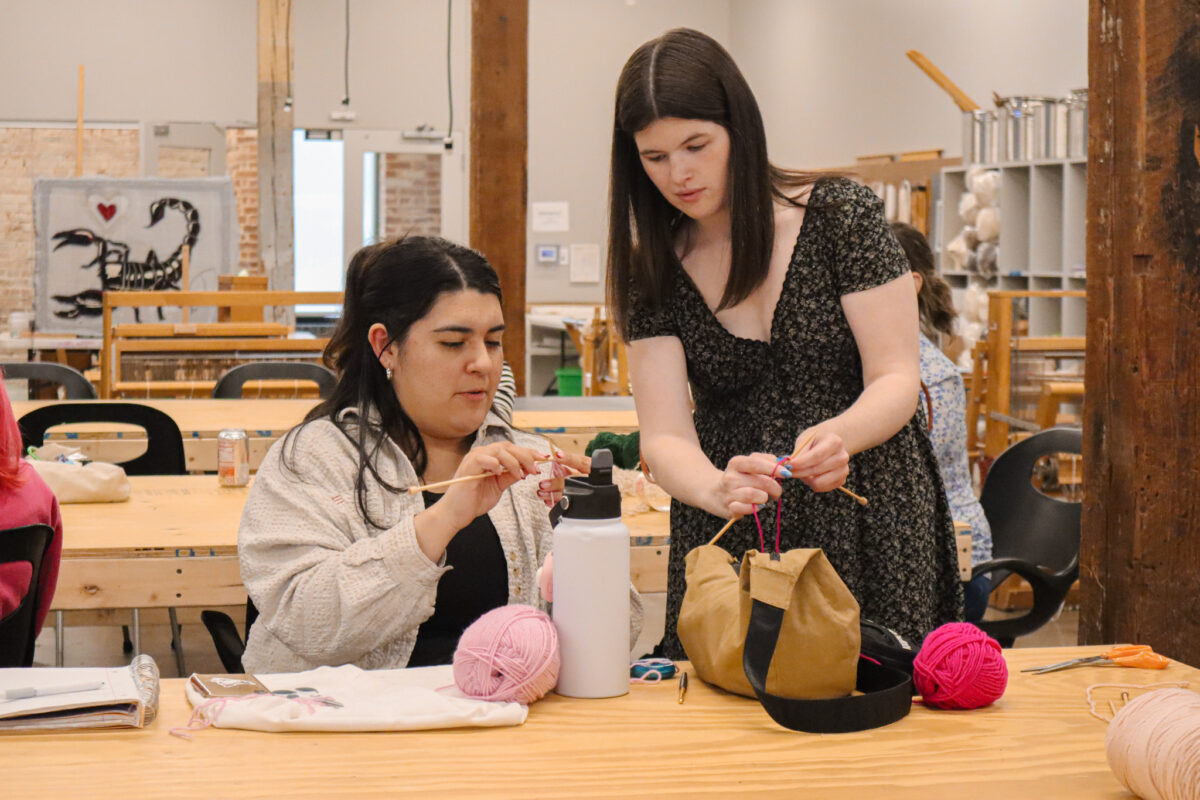A students sits at a table copying a knitting technique demonstrated by the instructor standing next to them
