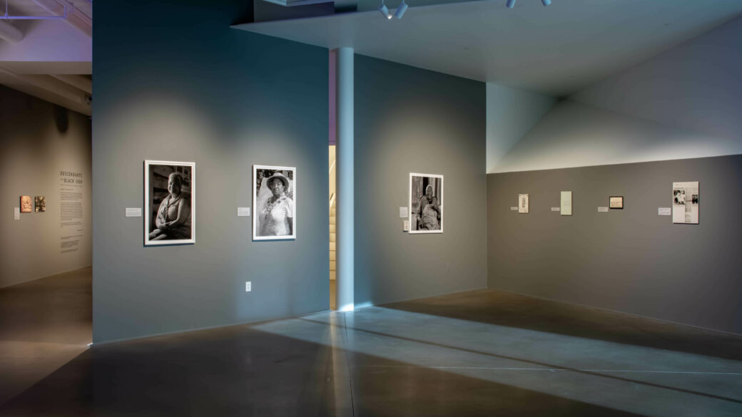 Black-and-white photographic portraits and facsimiles of historical photographs and newspapers hang in a gallery with gray walls