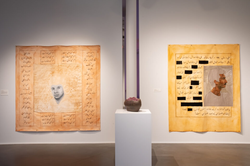 Two large canvases with portraits and Farsi writing hang behind a pedestal holding a ceramic bowl with dried flowers