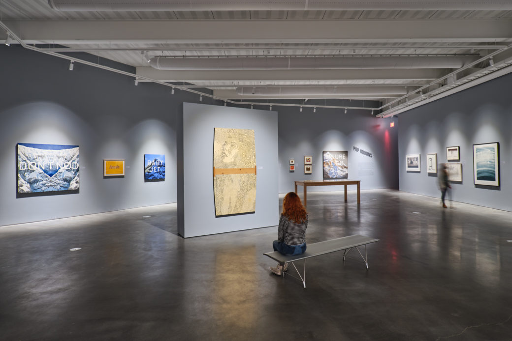 A person sits on a bench inside an art gallery with gray walls and numerous 2D works hung on the walls.