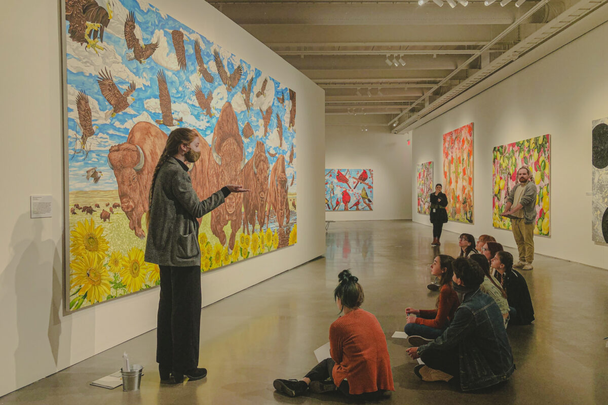 A group of students sits in front of a large painting of buffalo while a teacher addresses them