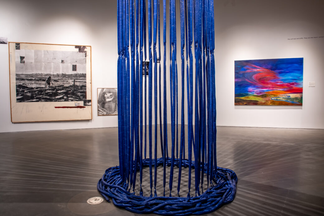 A collage of black-and-white photographs and a painting of a brightly colored sky and landscape hang on a wall. A column of blue tubes hang from the ceiling and pool on the floor in front of them.