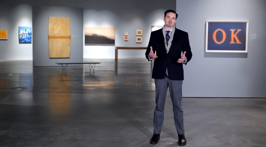 A person in a blazer and slacks stands gestures from inside an art gallery space