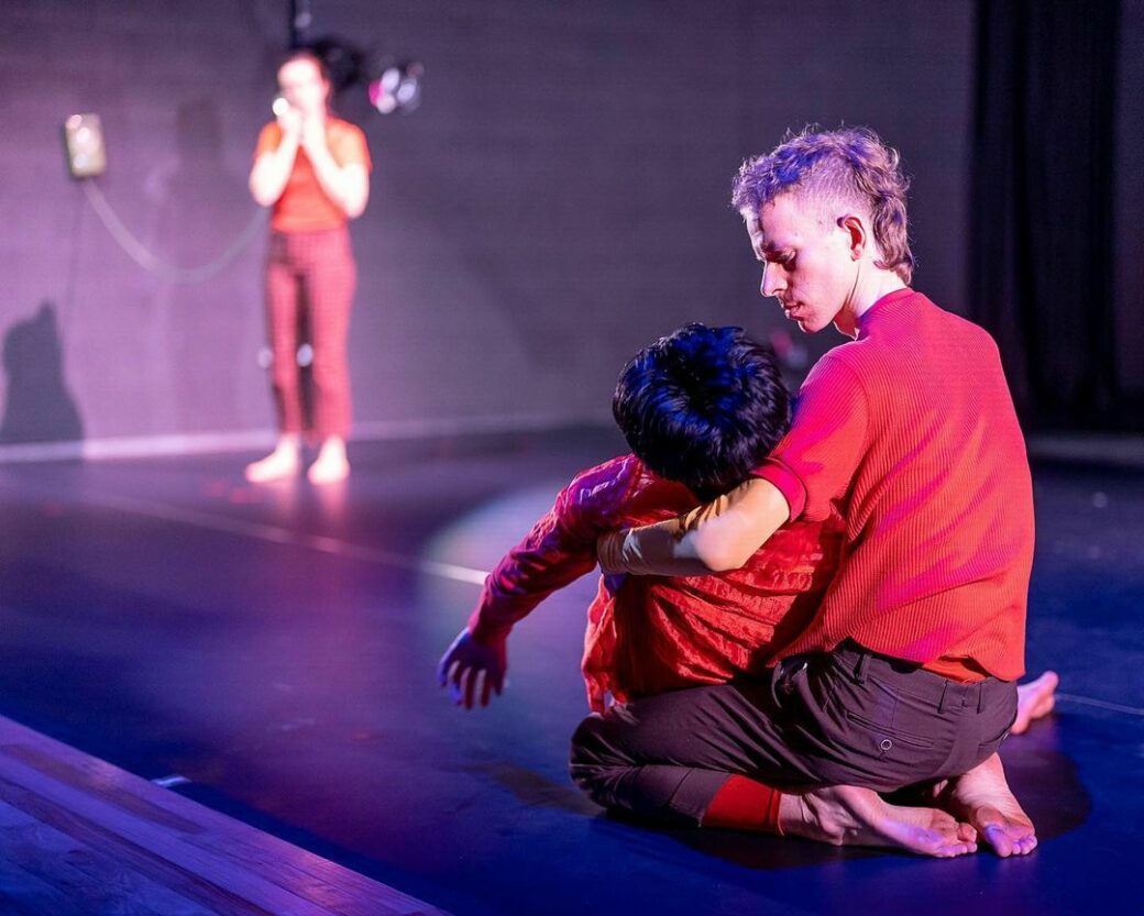 A performance view shows two people in the foreground. They are facing away from the camera while one holds the other in their arms like they have passed out. In the background we can see a person on a landline phone. Everyone is dressed in shades of red.