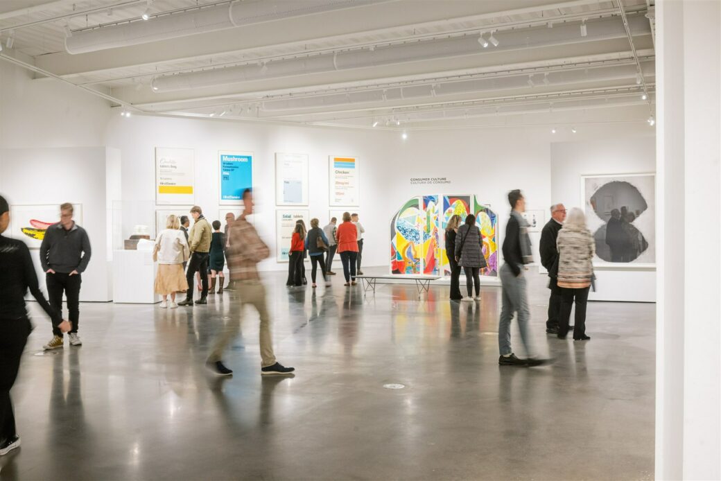 An art gallery is full of people, some standing while others walk, their movements captured in blur. Bright pieces of art hang on the walls and stand in the large room, with pops of blue, greens and yellows