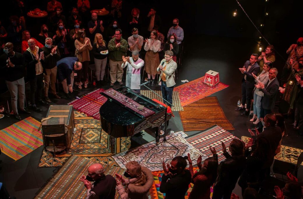 An audience is standing in a circle clapping their hands. In the center is an arrangement of colorful rectangle rugs. On top, a piano. Two people stand in the center, also clapping.