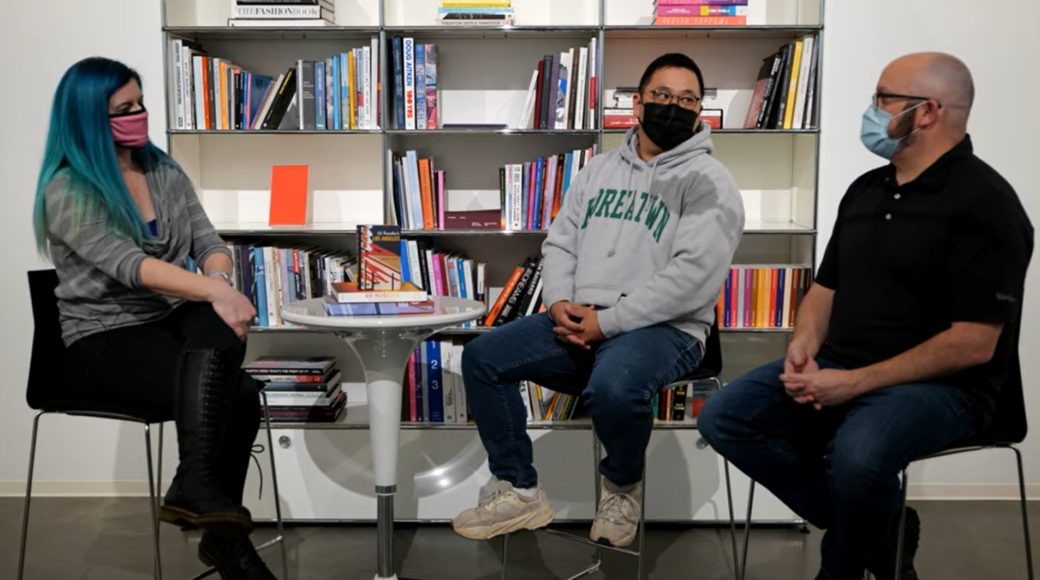 Three masked people sit around a circular table in front of a three-column book shelf
