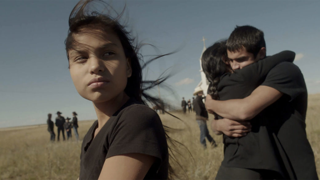 A film still depicts deep field of vision on an open prairie: a young girl stares into the distance in the foreground as a couple embraces behind her
