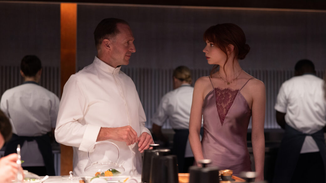Two people are standing in a restaurant kitchen, one a chef with a chef's coat, the other in a strappy pink dress. They are looking at one another, in conversation. Behind them we see other chefs working.