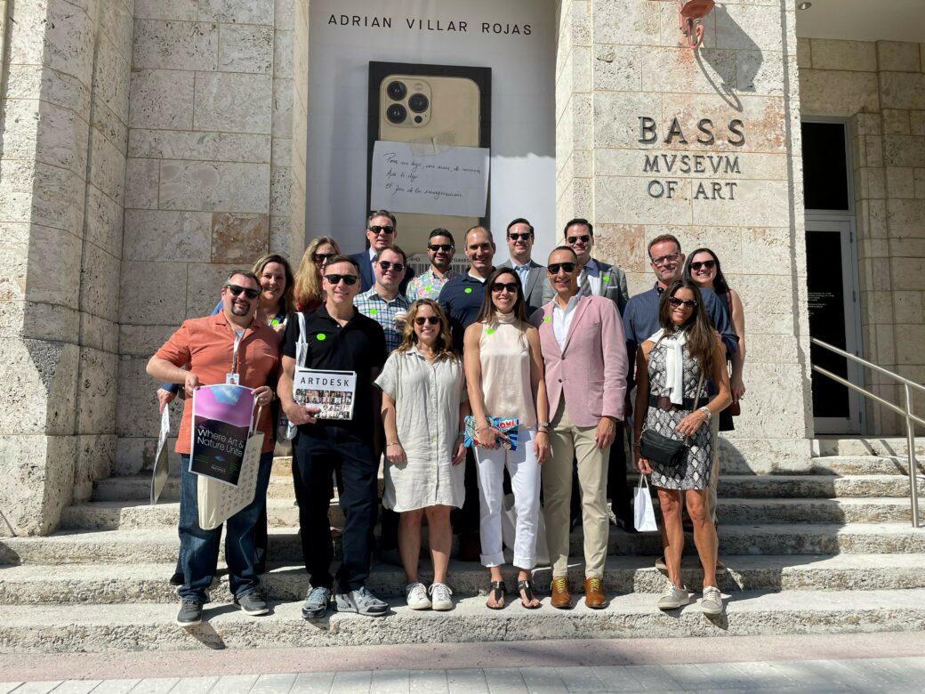 A group of people, many wearing sunglasses, stand on light-color stone steps in front of a matching building labeled "Bass Museum of Art"