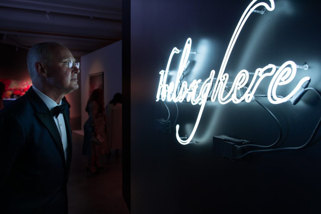 A man in a tuxedo looks at a glowing white neon sculpture with the text, “I belong here”