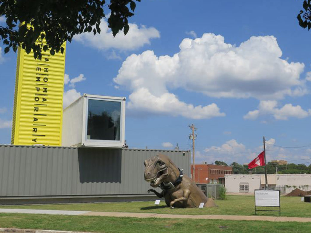 A dinosaur statue sits on a grass lawn in front of a gray building