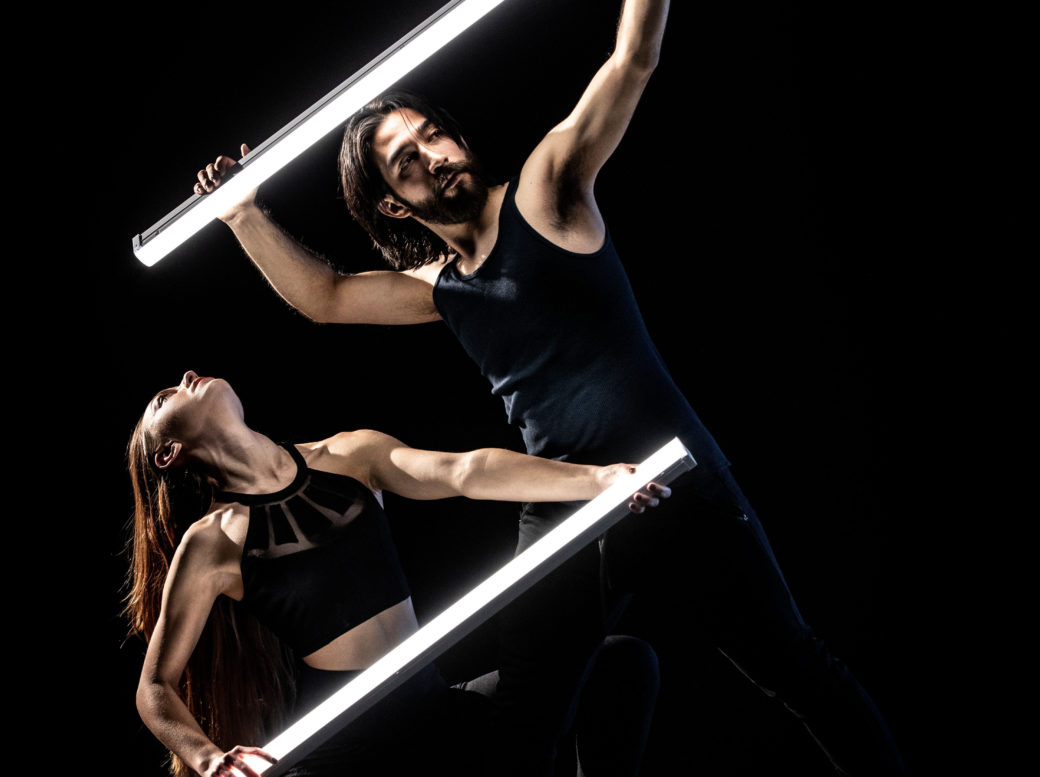 Two dancers pose with light sticks against a dark background
