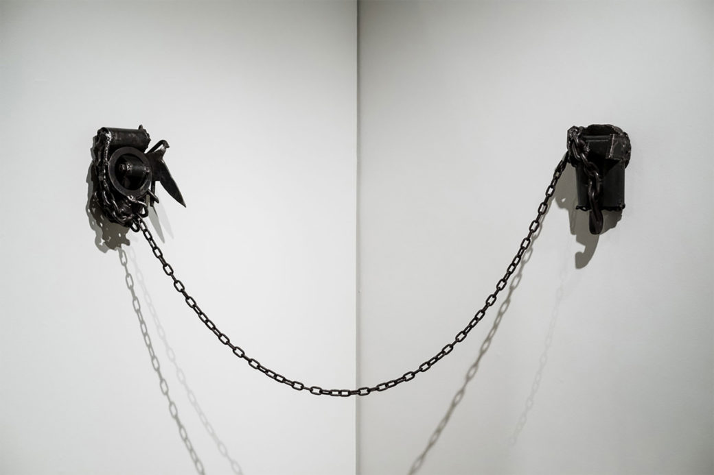 A sculpture of a black metal chain strung across two white walls that meet at the corner