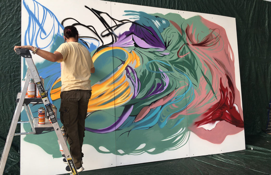 A person stands on a ladder with multiple cans of aerosol paint and actively paints a large-scale mural on three white panels