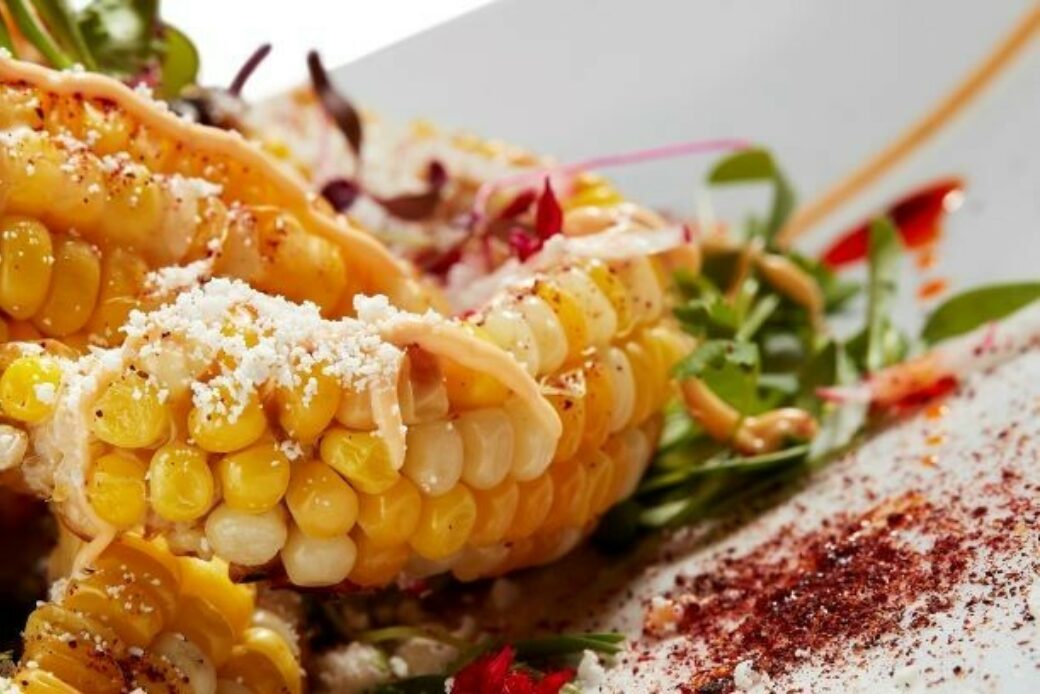 Grilled strips of corn are curled up on a white plate. We can see fresh greens and behind and on top. A red powder is sprinkled on top and on the plate.