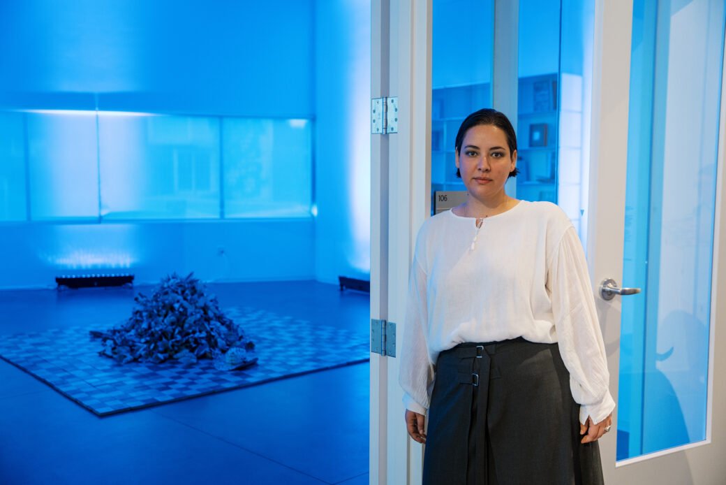 A woman stands in front of a glass door, leaning against the frame. Behind her is a blue-lit room, with a square of checkered tiles sitting in the middle. A pile of newspaper flowers sit on the tiles.