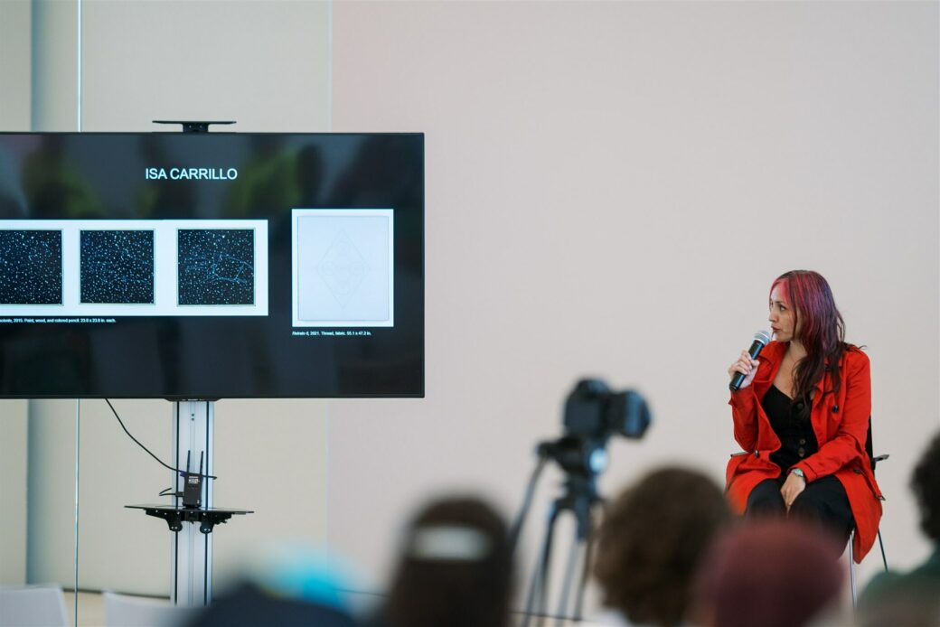 A person with long pink hair and a red coat over a black dress is sitting, turning to their right looking at a screen, talking into a microphone. On the black TV screen is the name ISA CARILLO and images of a constellation triptych