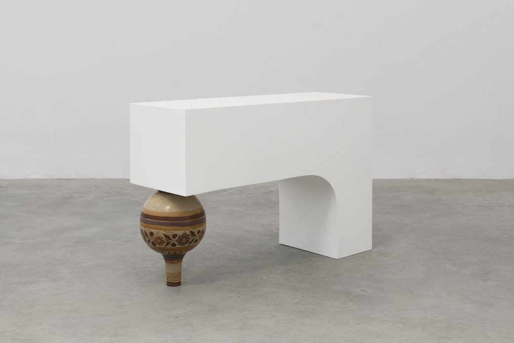 A white L-shaped block rests on its short leg with the long leg supported by an upside down vase