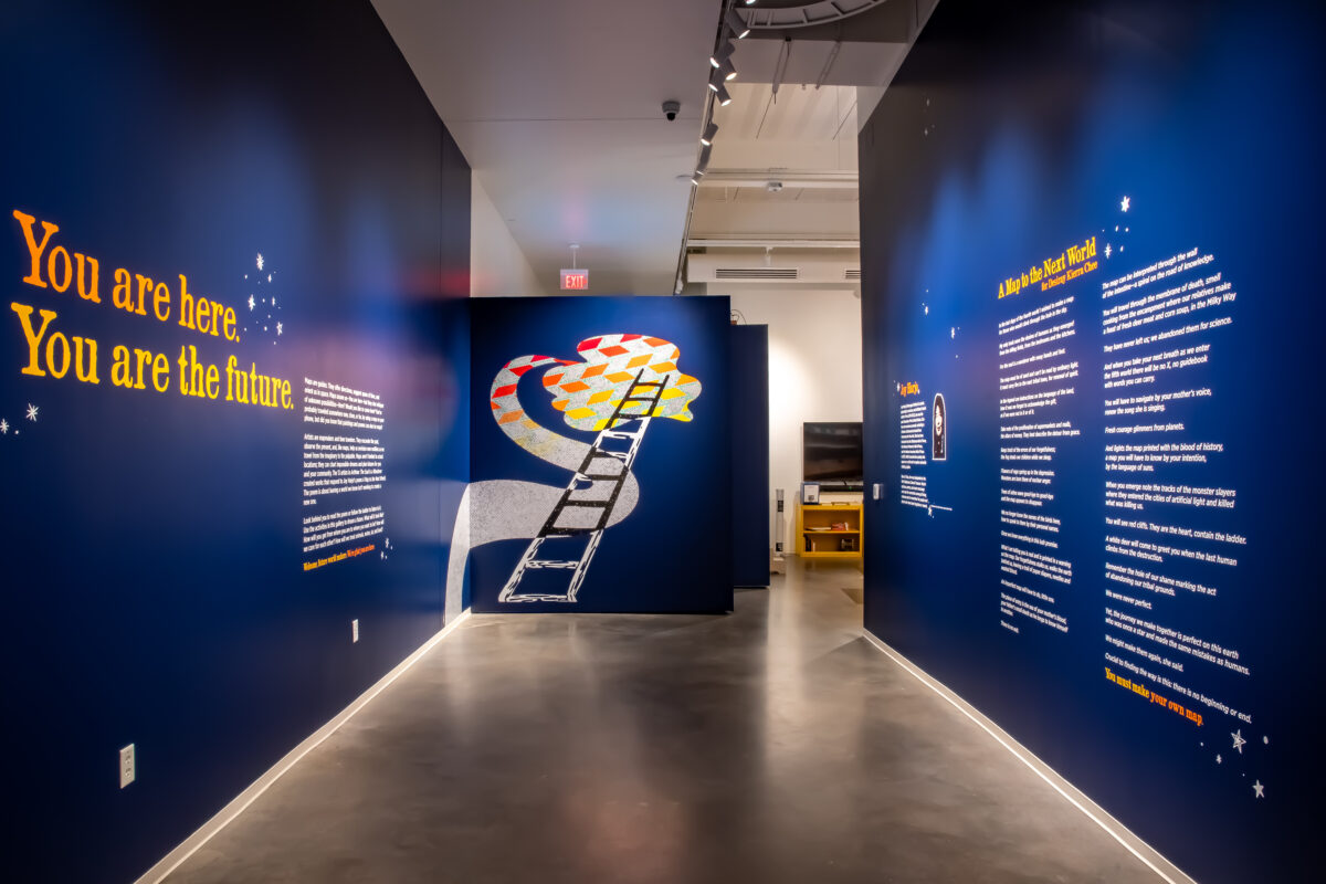Dark blue walls on either side. The left wall says "You are here. You are the future." An image of a colorful ladder is at the back. To the right, white and yellow text is on the blue wall.