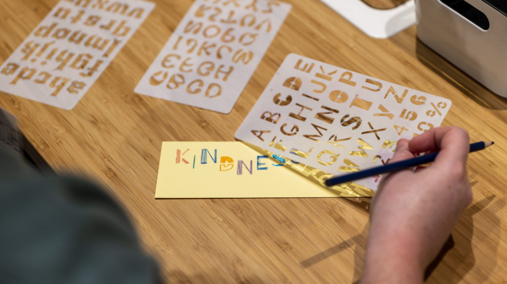 A person uses a set of stencils to transfer the word "kindness" onto a bookmark-sized piece of cream-colored paper