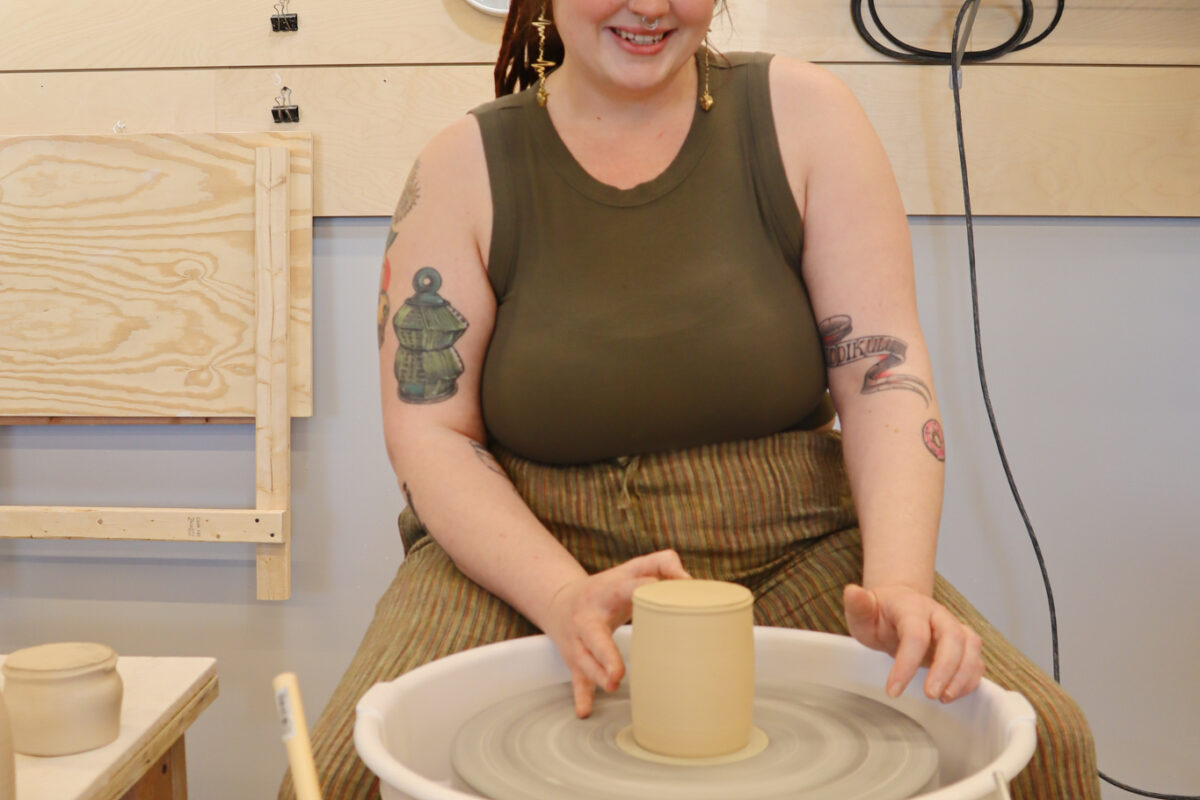 Woman with long brown hair pulled back smiles while centering a pot on the potters wheel