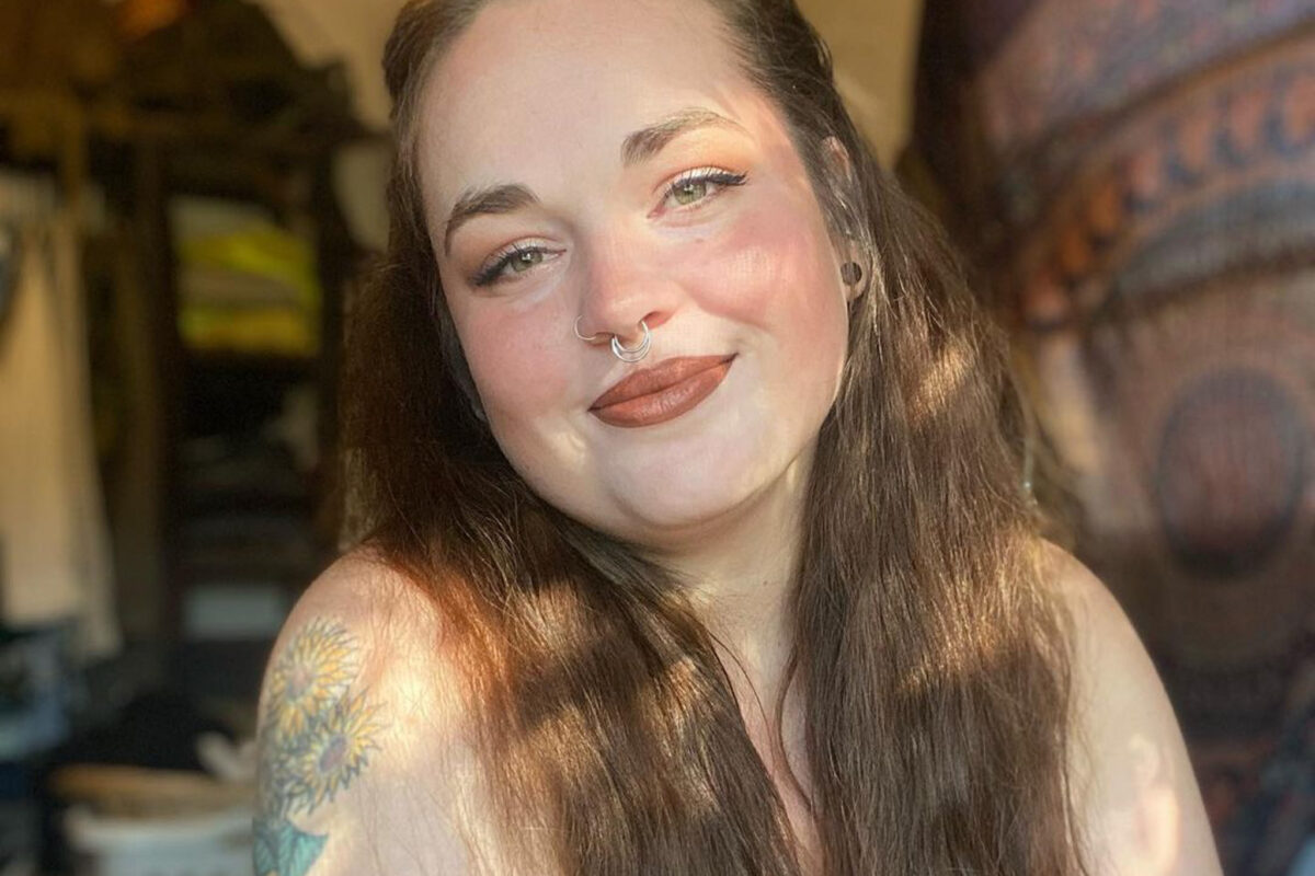 A white woman with long brown and a septum ring hair smiles softly at the camera. We can see colorful tattoos on her right arm.