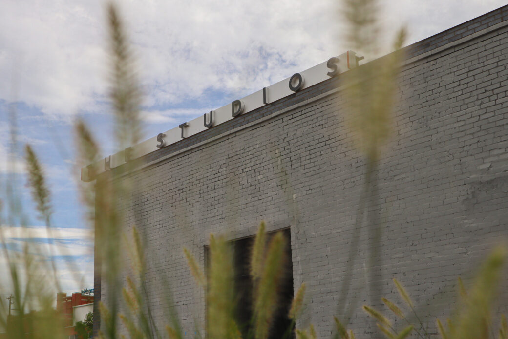 A gray-bricked building stands against a cloudy blue sky. A sign at the top reads THE STUDIOS. Reeds of grass blur the image.