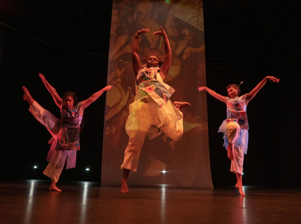 Three dancers in flowing costumes perform in front of a scrim in a dark theater